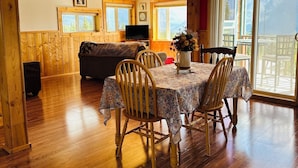 A wooden cabin interior with a dining table, chairs, a sofa, and a TV.