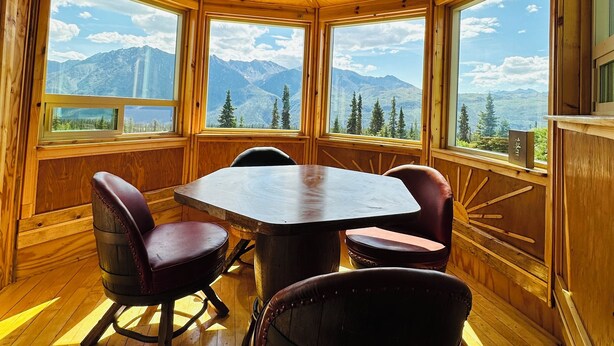 A wooden interior with a large table and chairs, large windows with mountain views, and a wooden ceiling.