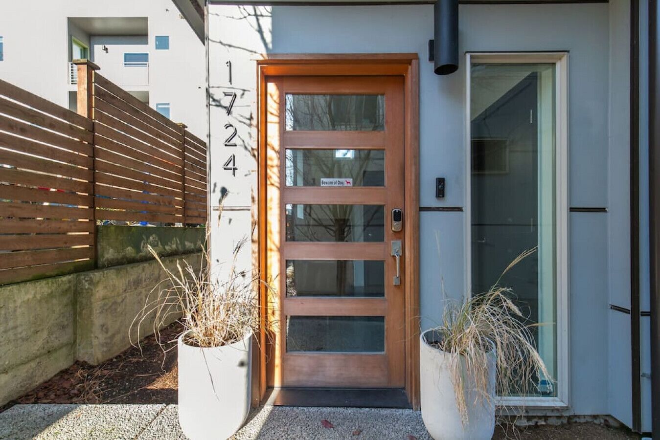 A modern building entrance with a wooden door, numbered 1724, and two potted plants.