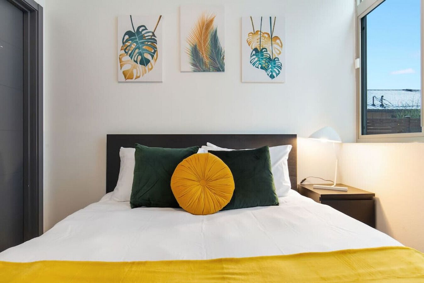 Een net opgemaakt bed met wit en geel beddengoed, groene en gele kussens, en drie kunstwerken met bladmotieven erboven.