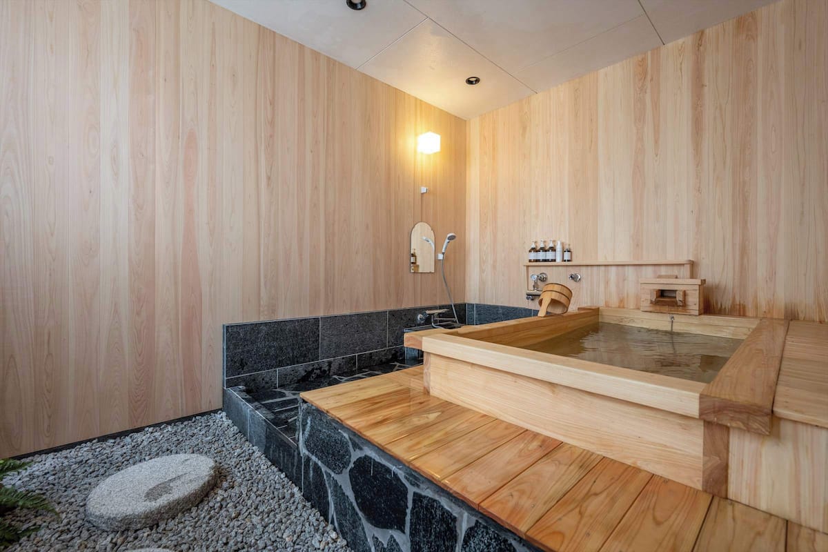 Une salle de spa en bois, avec une baignoire en bois, un lavabo en pierre et une étagère en bois où sont posées des bouteilles.