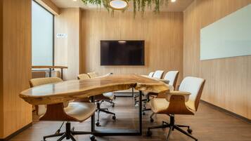 A conference room with a large wooden table, white chairs, a mounted TV, and a hanging plant.