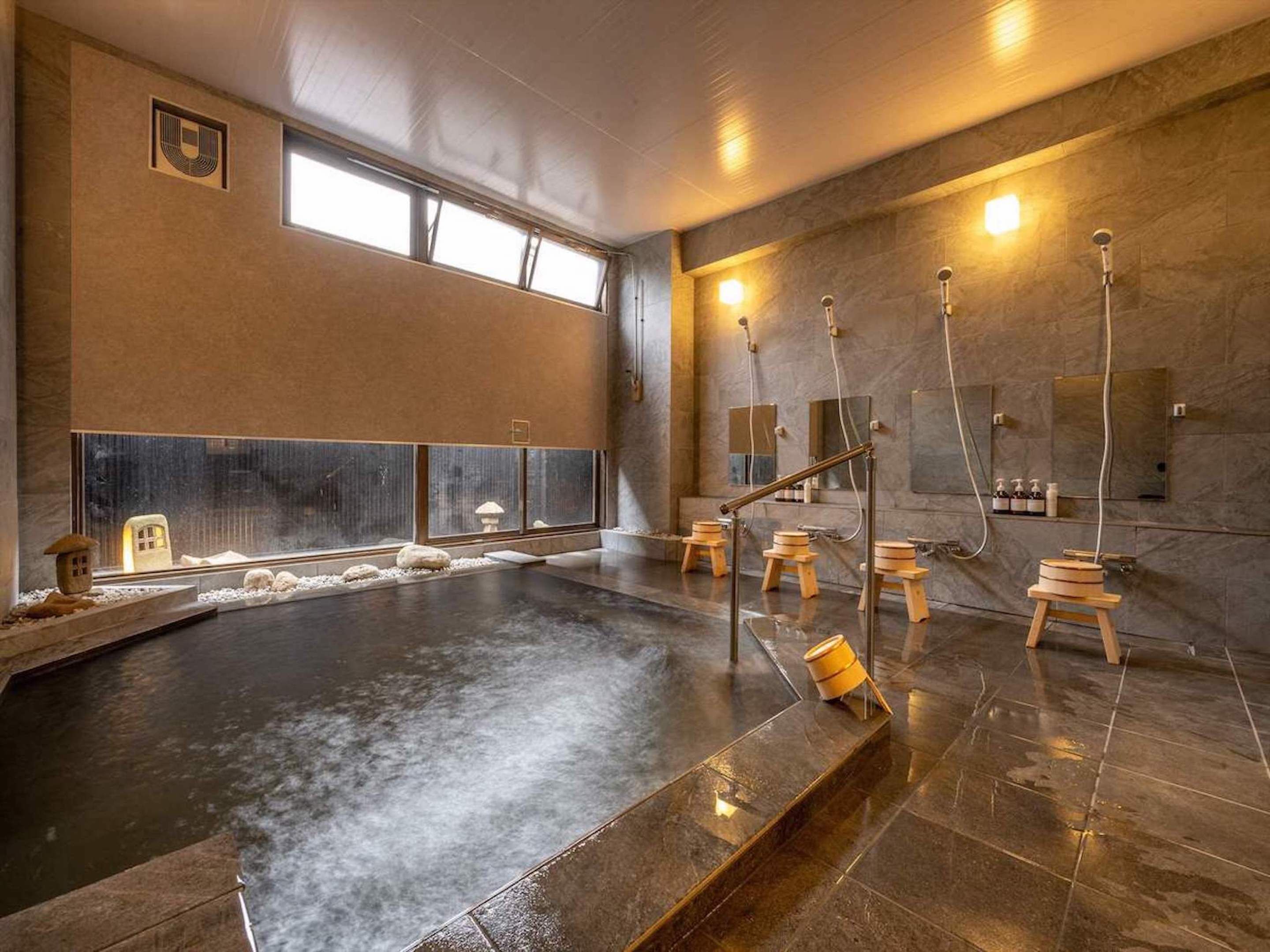 Une piscine intérieure moderne avec un sol en pierre, des tabourets en bois et un mur en pierre agrémenté d’une fenêtre.
