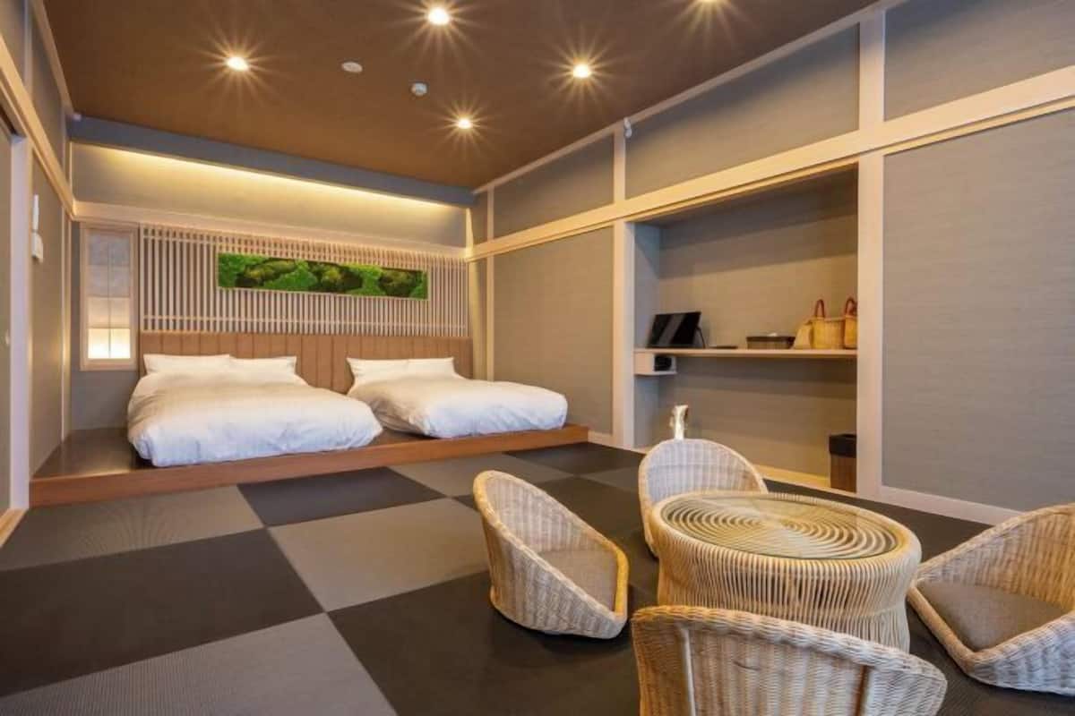 Ein Hotelzimmer mit zwei Betten, einer Sitzecke mit Korbstühlen, einem an der Wand befestigten Regal mit Gegenständen und einem großen Fenster.