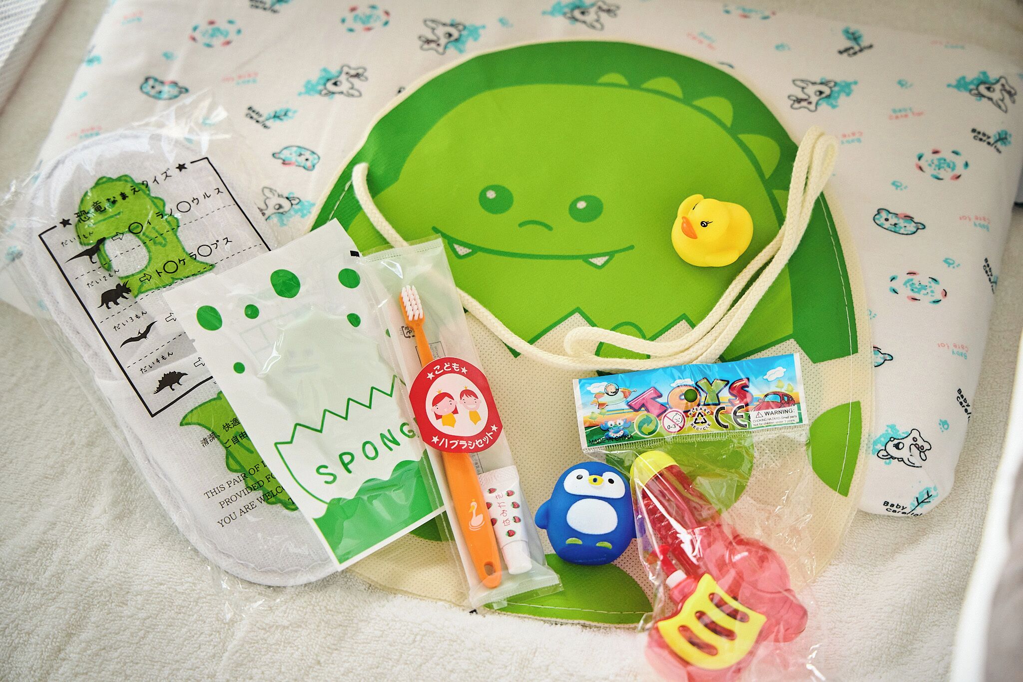 A collection of baby items including a green plush toy, a yellow rubber duck, a blue toy, and a pink toy.