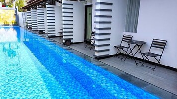 Una piscina con borde de azulejos, junto a un edificio con columnas blancas y una puerta. Hay dos mesas y cuatro sillas afuera del edificio.