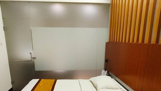 A single bed with white bedding and a brown accent, a wooden headboard, a bedside table, and a wardrobe with a mirror. - Odaita Hotel (Pamekasan)