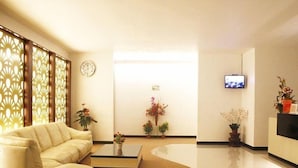 A modern hotel lobby with a curved sofa, a coffee table, decorative plants, a clock, and a television on the wall. - Odaita Hotel (Pamekasan)