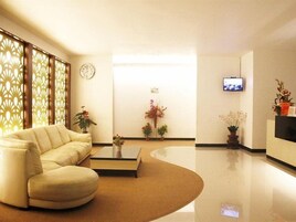 Un moderno hall de hotel con un sofá curvo, una mesa de centro, plantas decorativas, un reloj y un televisor en la pared.