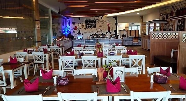 A restaurant with white chairs and tables, a bar area in the background, and a wall with various decorations. - Odaita Hotel (Pamekasan)