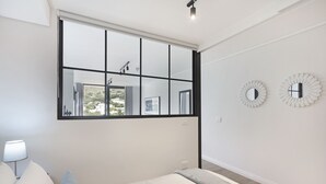 Un dormitorio moderno con un ventanal grande, una cama con ropa de cama blanca, una lámpara de noche y dos espejos fijados a la pared.