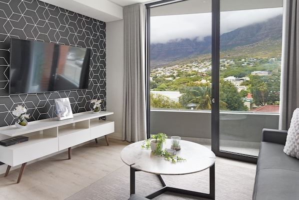 Un salon moderne doté d’un grand téléviseur, d’une console blanche, d’une table basse ronde agrémentée de plantes et d’un balcon offrant une vue sur la ville.