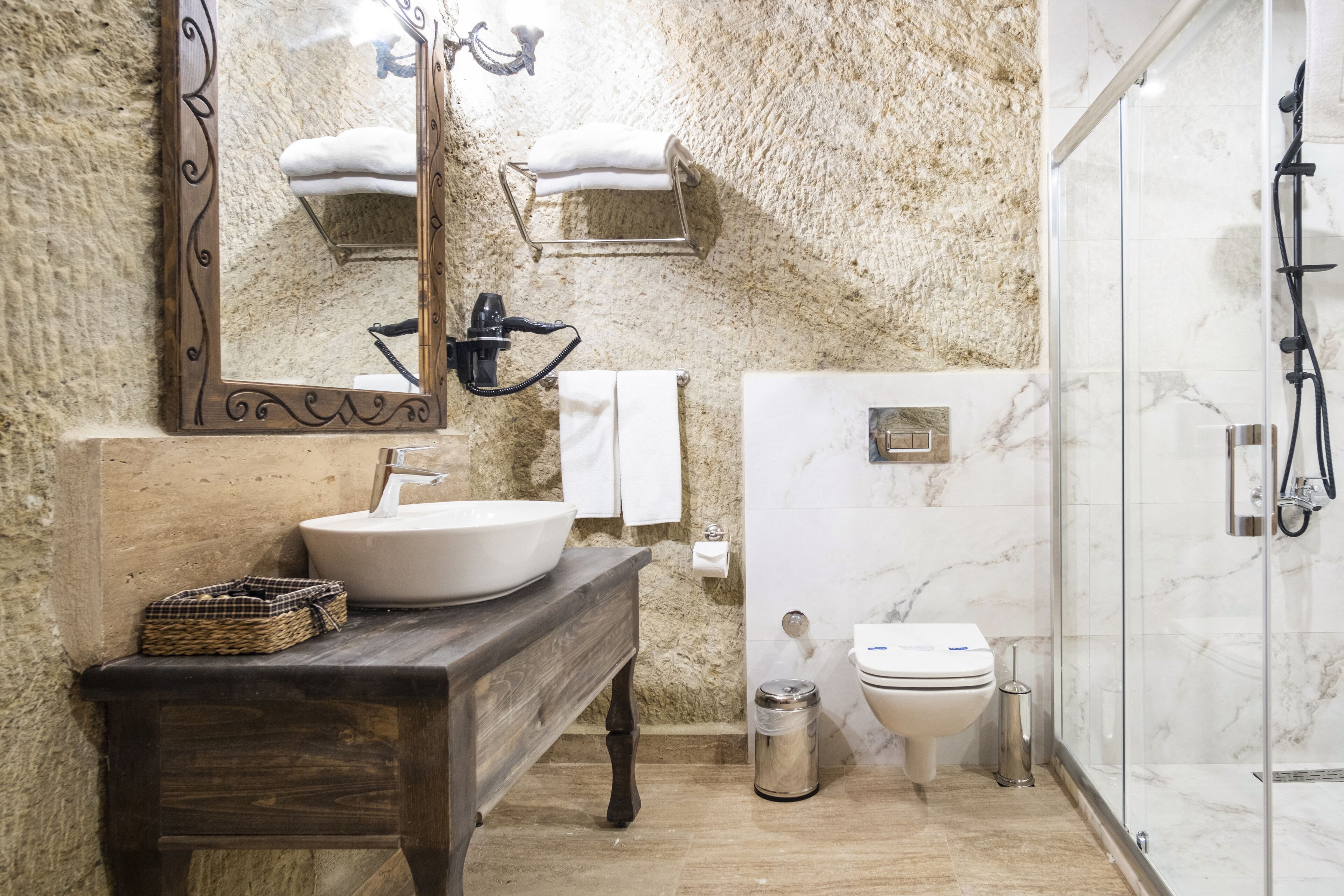 A bathroom with a marble wall, a wooden vanity, a large mirror, a sink, a toilet, and a glass-enclosed shower.