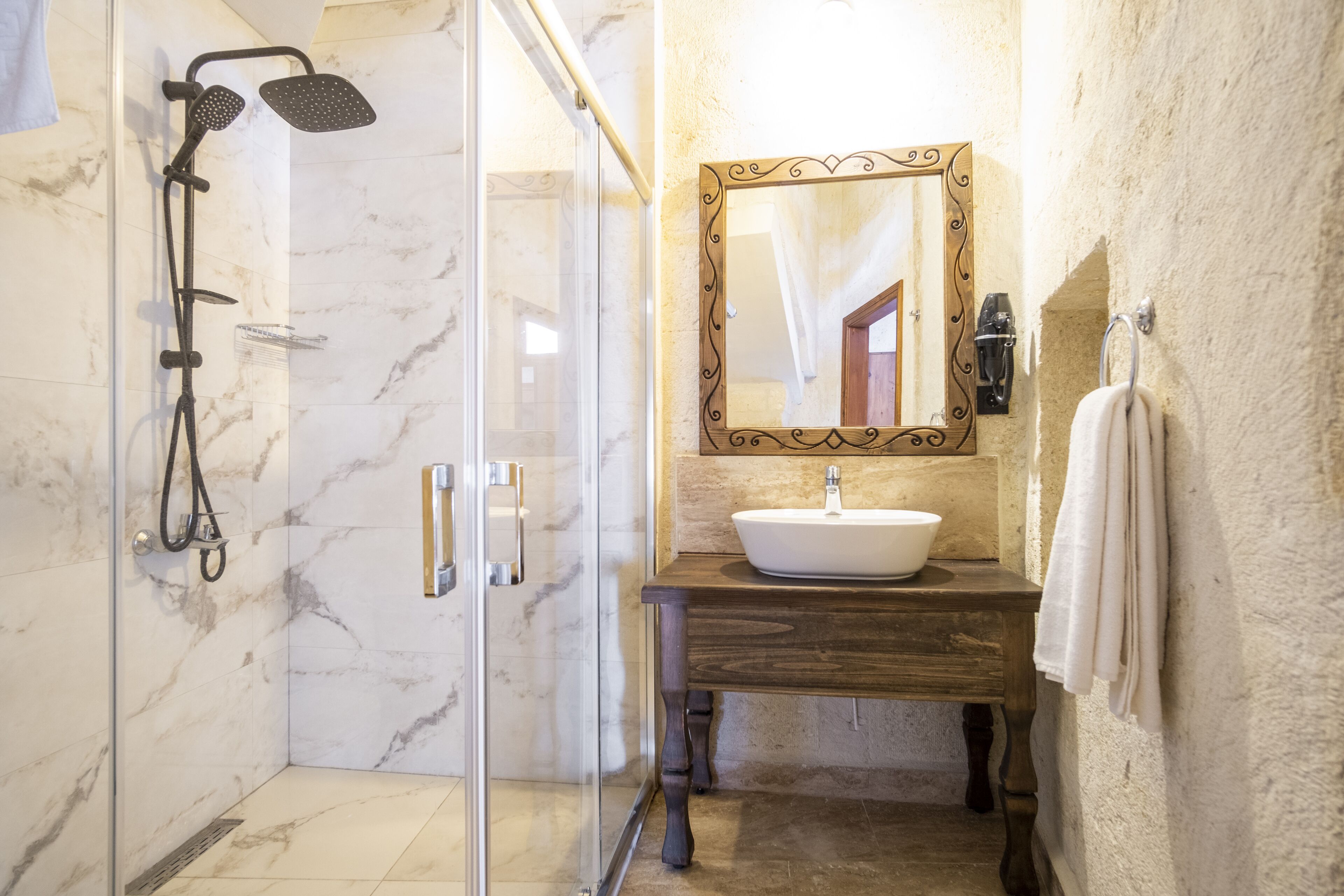 Un baño con ducha envuelta en vidrio, tocador de madera con lavamanos blanco y espejo con marco decorativo.