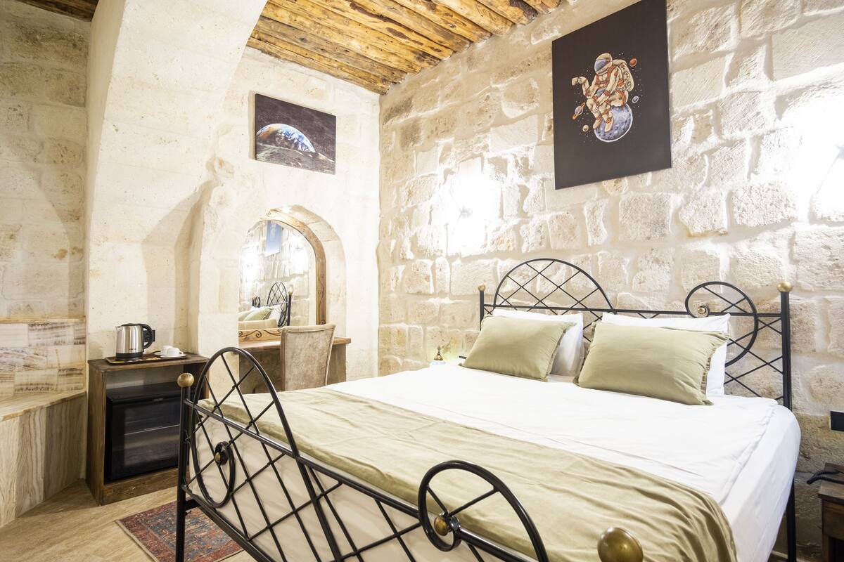 Un dormitorio con pared de piedra, cama con estructura metálica, mesita de noche de madera y una pequeña mesa con tetera.
