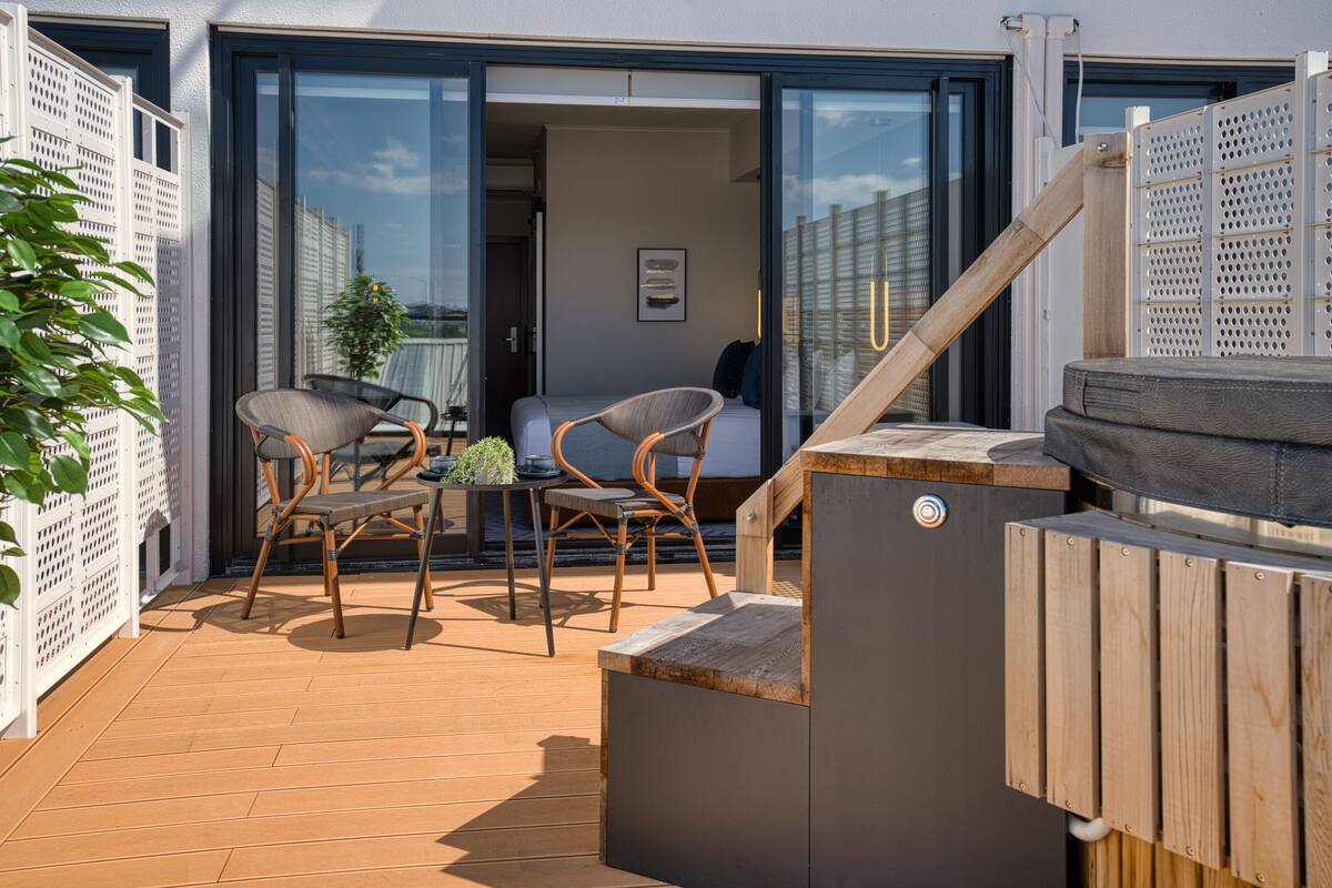 A balcony with a wooden deck, two wicker chairs, a small table, and a view of the interior through a glass door.