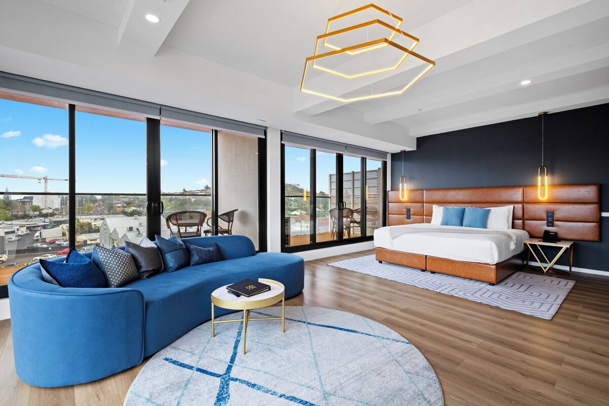 A modern bedroom with a large bed, a blue sofa, and a city view through large windows.