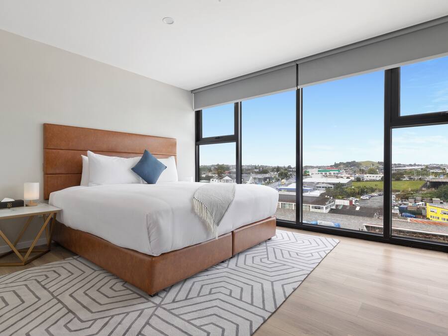 A modern bedroom with a large bed, a nightstand, and a view of a cityscape through large windows.