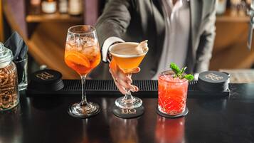 Drei Cocktails auf einer Bar, ein Barkeeper ist im Hintergrund zu sehen.