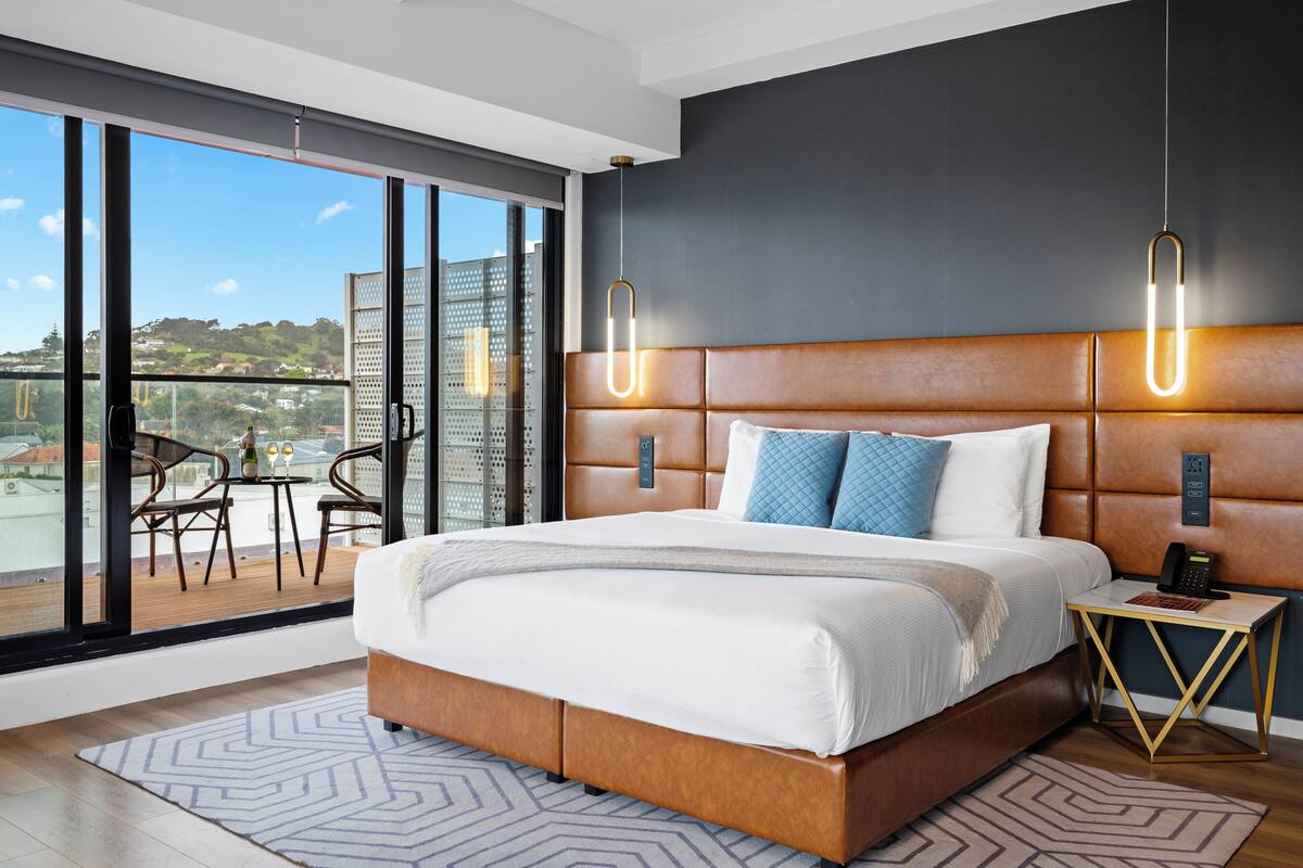 A modern hotel room with a large bed, a small table, and a view of the city through large windows.