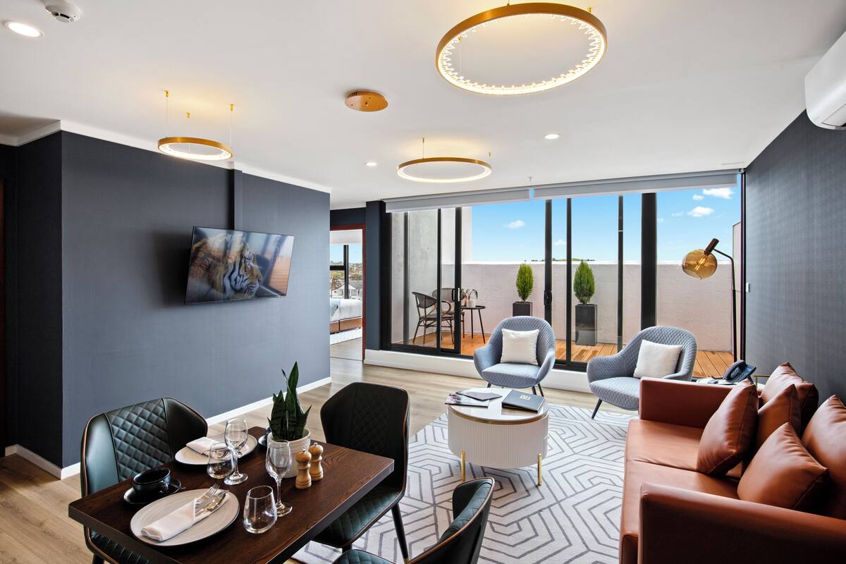 A modern dining area with a wooden table set for two, a television mounted on the wall, and a large window offering a view of the outdoors.