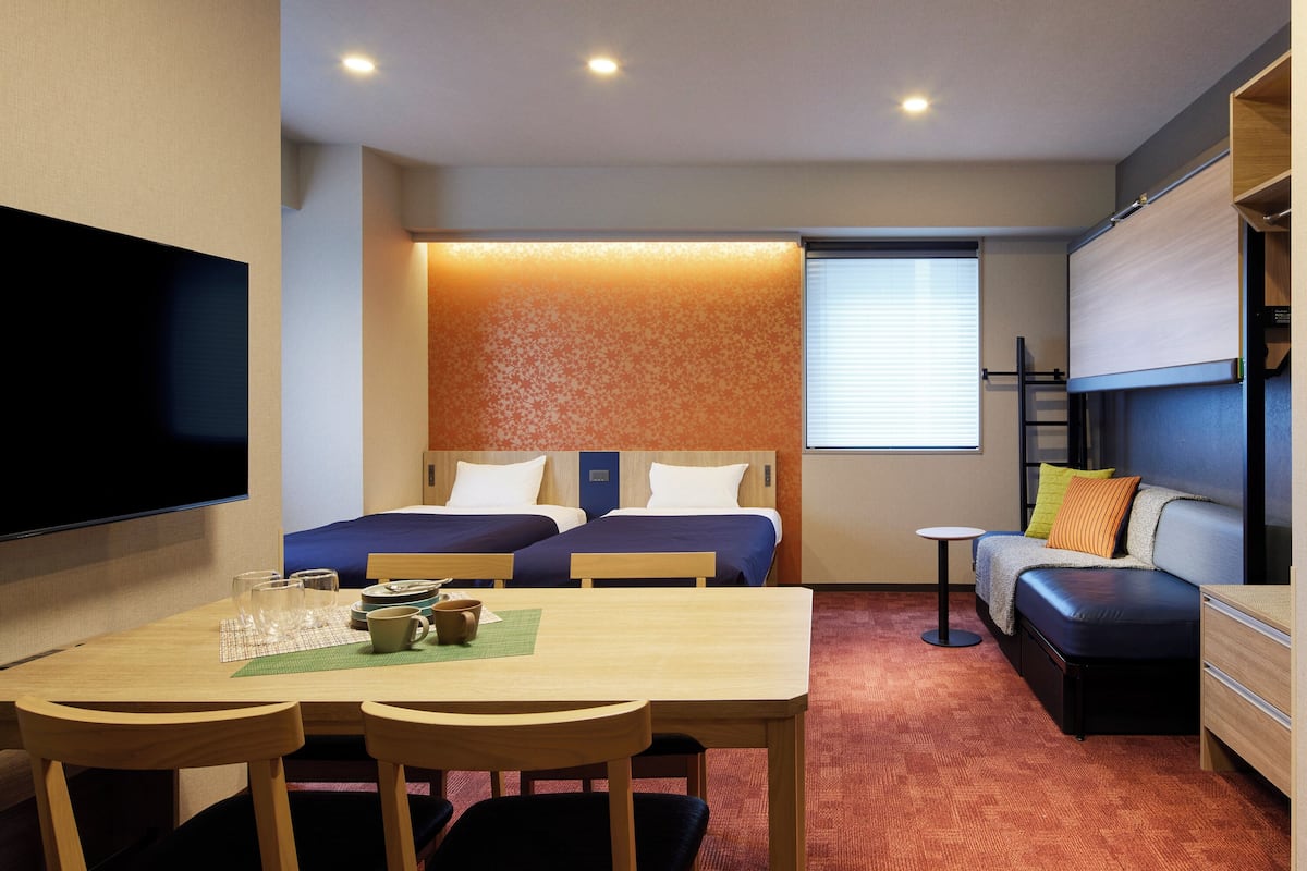 A compact hotel room with two beds, a sofa, a dining table with chairs, and a flat-screen TV.