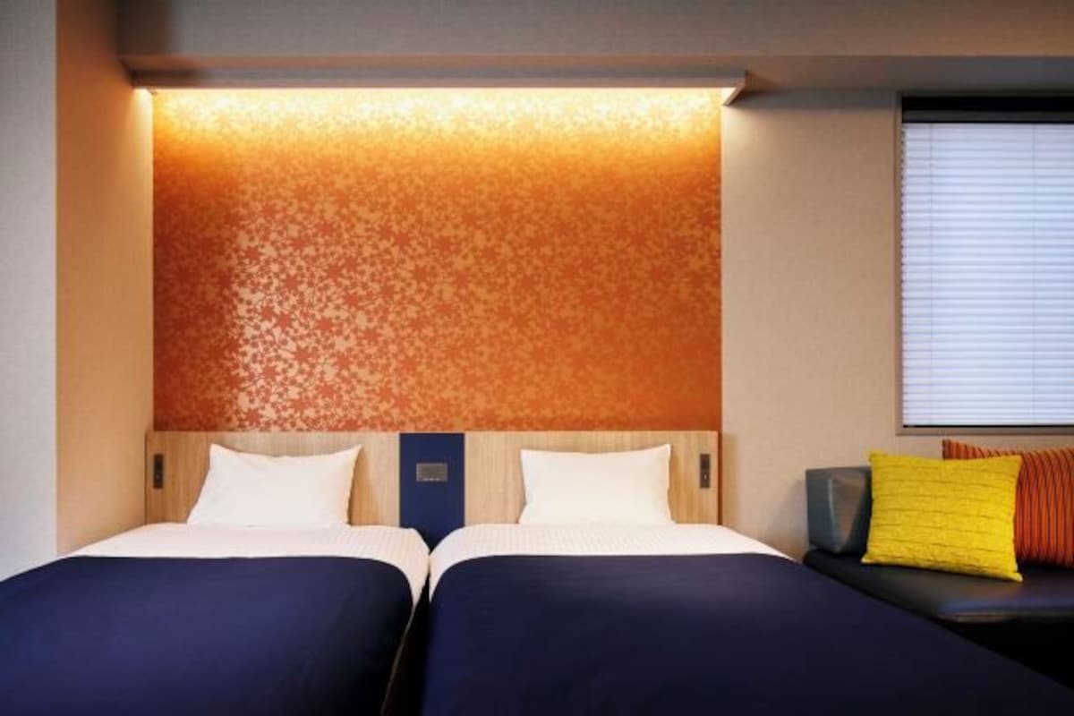 A hotel room with two beds, a headboard with a textured design, a window with blinds, and a bench with cushions.
