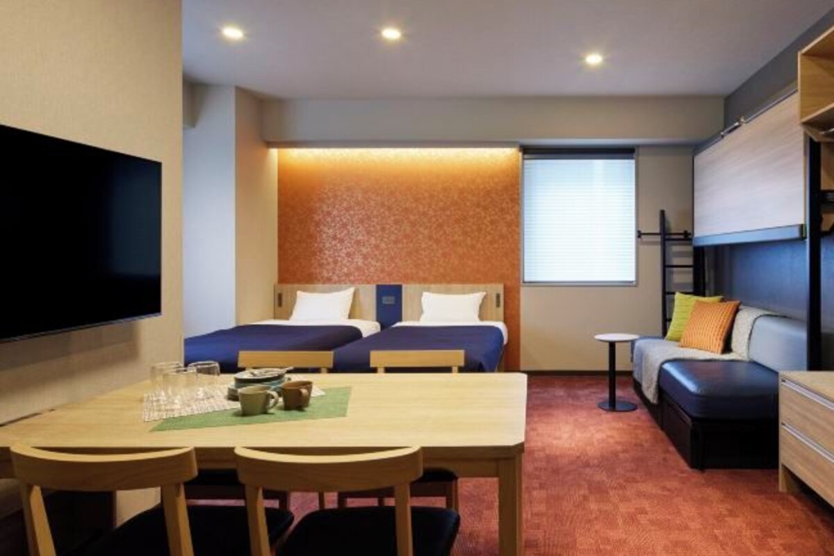 A compact hotel room with a bed, a sofa, a dining table with chairs, and a TV mounted on the wall.