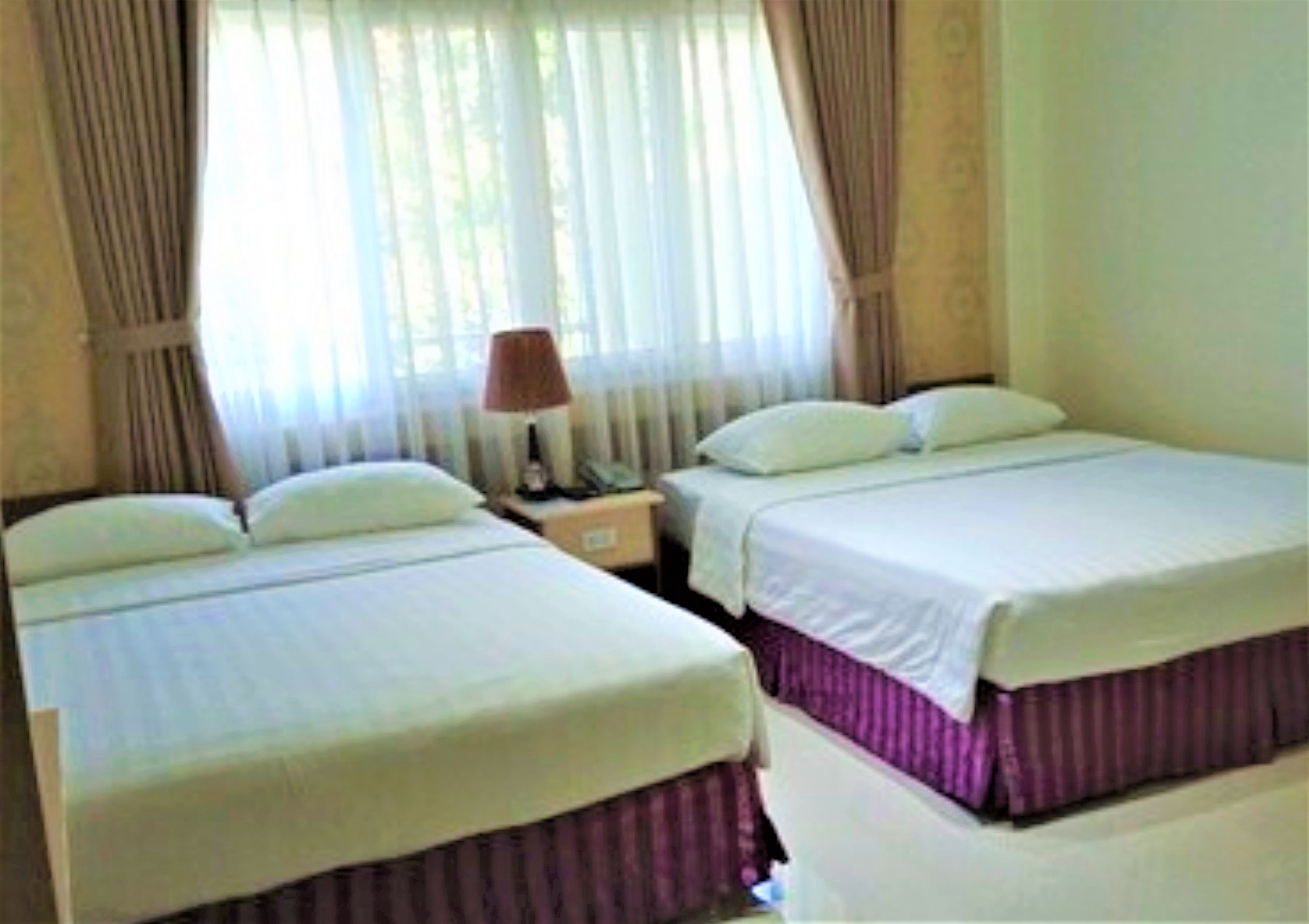 A hotel room with two beds, a nightstand with a lamp, and sheer curtains.