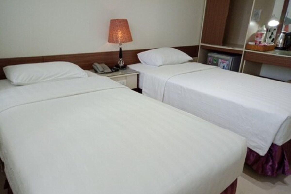 Quarto de hotel com duas camas de solteiro, uma mesa de cabeceira com abajur, telefone e uma prateleira pequena com objetos.