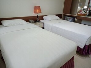 Quarto de hotel com duas camas de solteiro, uma mesa de cabeceira com abajur, telefone e uma prateleira pequena com objetos.