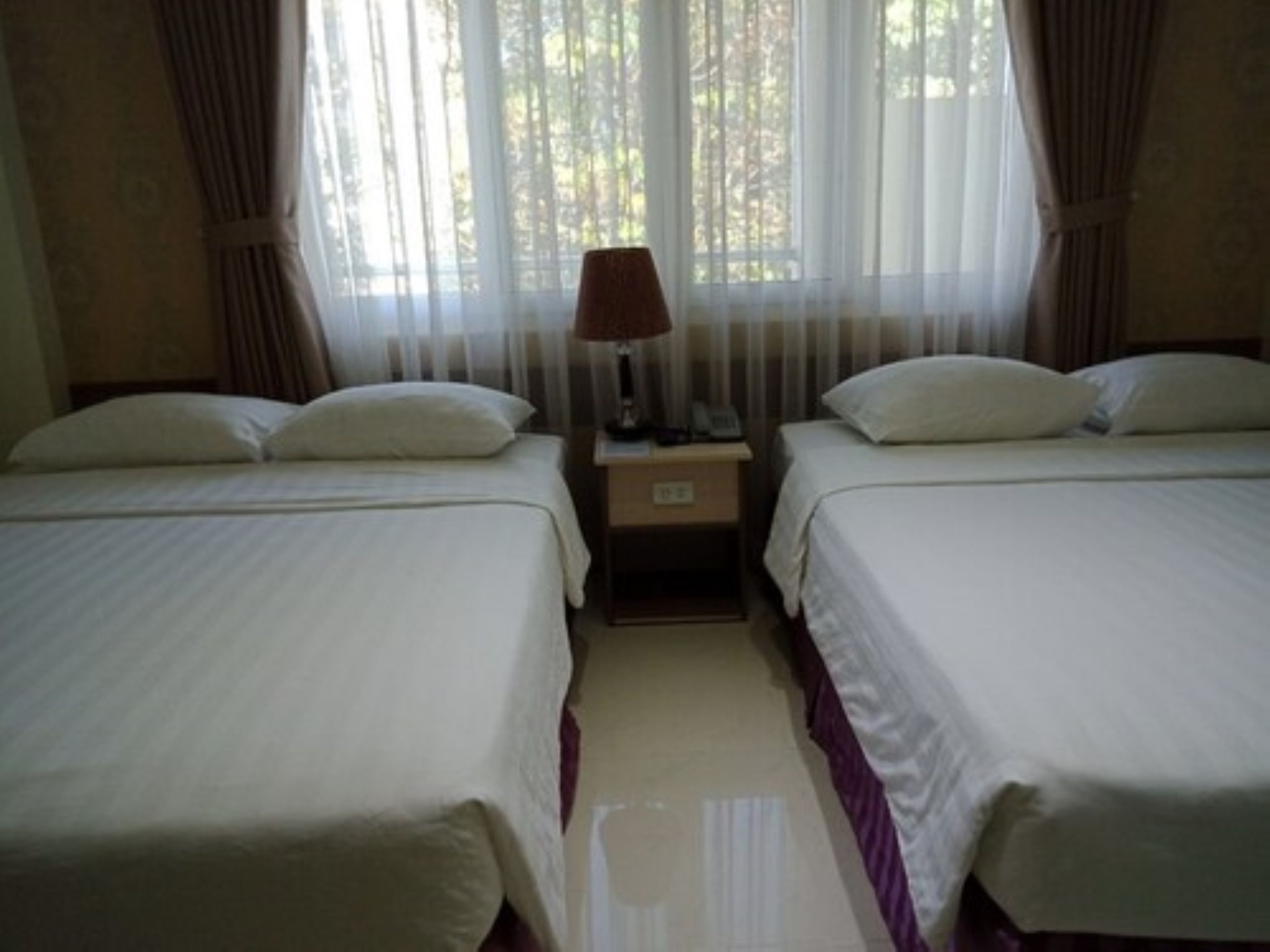 Une chambre d’hôtel avec deux lits, une table de chevet, une lampe et une fenêtre avec des rideaux légers.