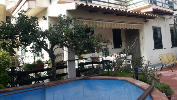A two-story house with a balcony, a small pool, and a garden with trees and plants.