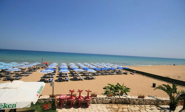 A beach with rows of blue and white umbrellas, a stone wall with potted plants, and a sign for Heineken.