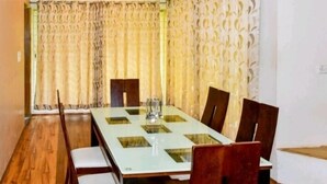 A dining area with a rectangular glass table surrounded by wooden chairs, beige curtains, and patterned wallpaper. - Surya Kiran Miramar (Panaji)