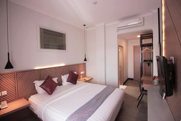 Una habitación de hotel con una cama bien hecha, una mesita de noche con lámpara, un televisor montado y un escritorio pequeño con una silla.