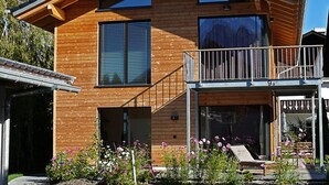 A modern two-story wooden house with a balcony, large windows, and a small garden with flowers. - Exclusive Mondholz vacation apartment in a very quiet and central location in Garmisch (Garmisch-Partenkirchen)