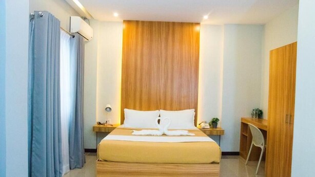 A hotel room with a wooden headboard, a bed with white linens, a desk with a chair, and a wardrobe. - ELTI Suites (Angeles City)