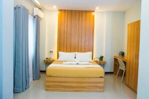 A hotel room with a wooden headboard, a bed with white linens, a desk with a chair, and a wardrobe. - ELTI Suites (Angeles City)