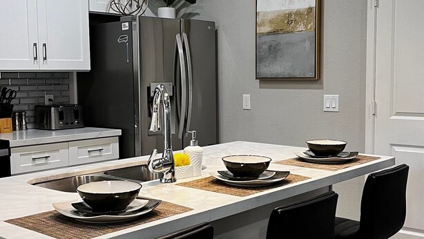 A modern kitchen with a central island, bar stools, stainless steel appliances, pendant lighting, and a minimalist design. - Cozy home near Medical Center & NRG Stadium - B (Houston)