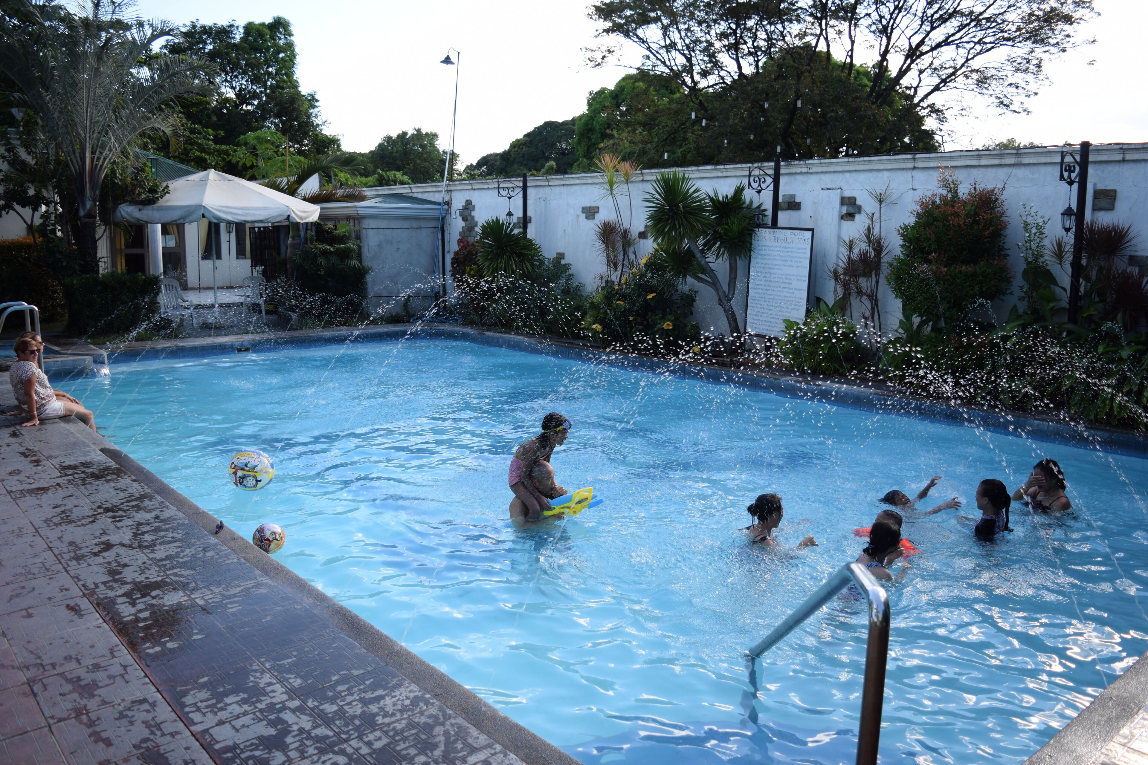 A swimming pool with children playing, surrounded by a white wall and greenery.