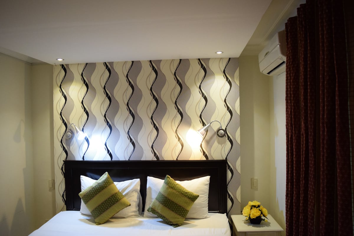 A neatly made bed with two pillows, a bedside table with a vase of yellow flowers, and a wall with a wavy pattern.