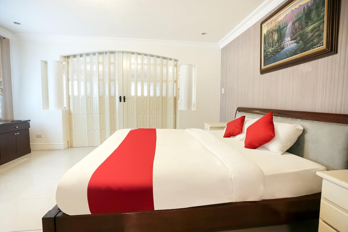 A neatly made bed with a red and white bedspread, a wooden headboard, and two red pillows. There is a painting of a landscape on the wall above the bed.