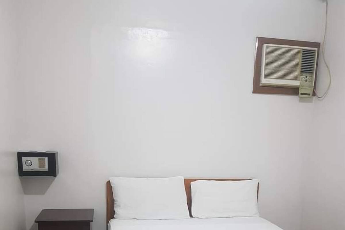 Quarto limpo e simples com cama de madeira, roupa de cama branca, uma pequena mesa de cabeceira e ar condicionado fixado na parede.