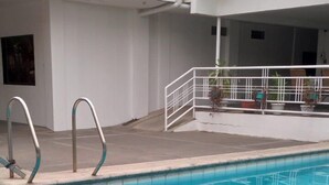 A swimming pool with metal ladders, a white building with windows, and a covered area with chairs and potted plants. - Platinum Hotel (Angeles City)