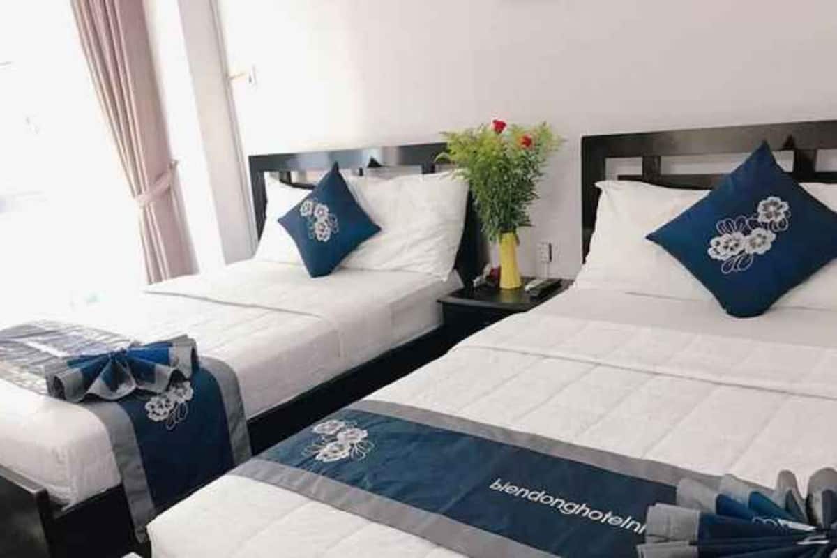 Ein Hotelzimmer mit zwei Betten, jedes mit einem blauen, floralen Kissen und einer passenden Tagesdecke. Ein kleiner Tisch mit einem Blumenstrauß steht zwischen den Betten.