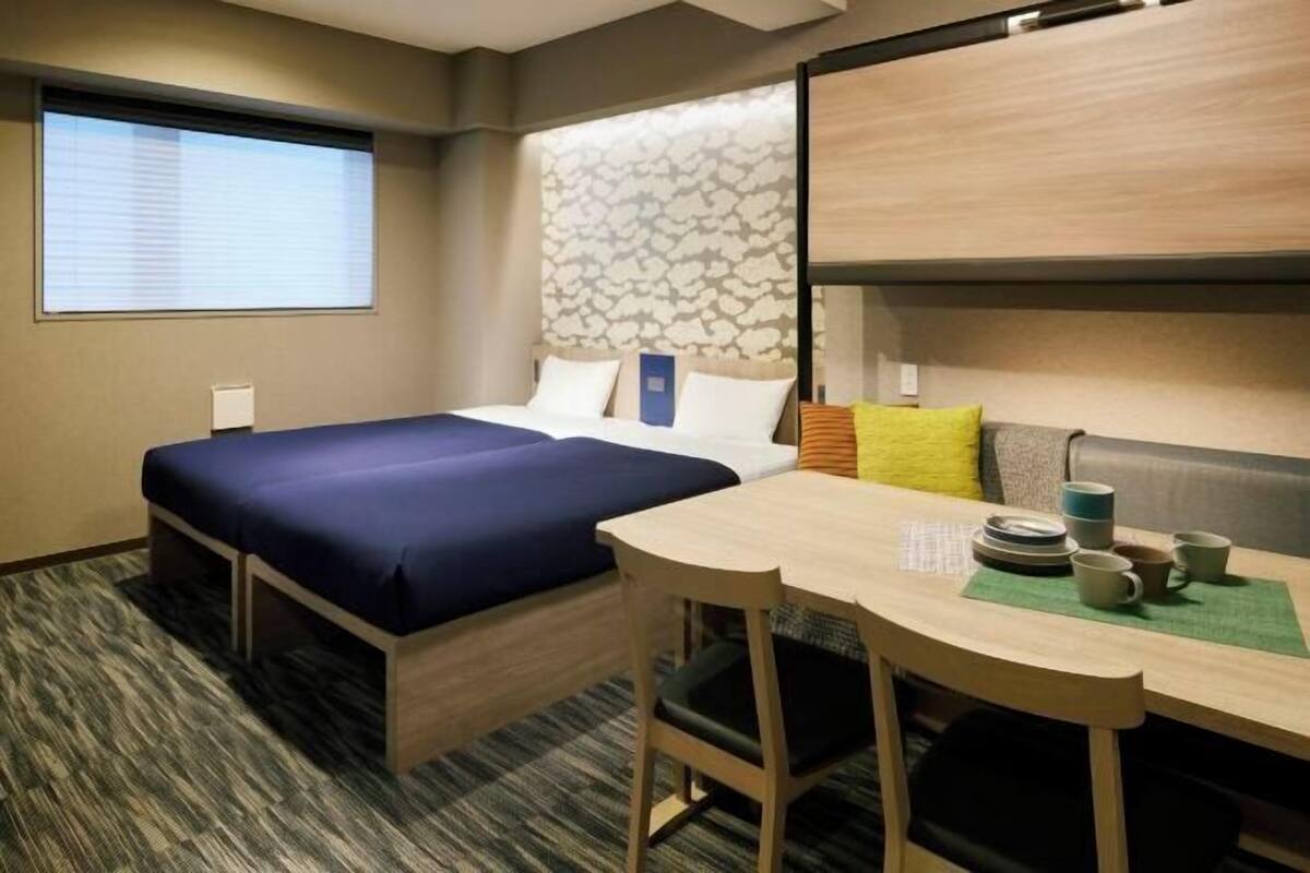 A hotel room with a bed, a desk, and a chair. There is a window with blinds, a wall with a leaf pattern, and a carpeted floor.