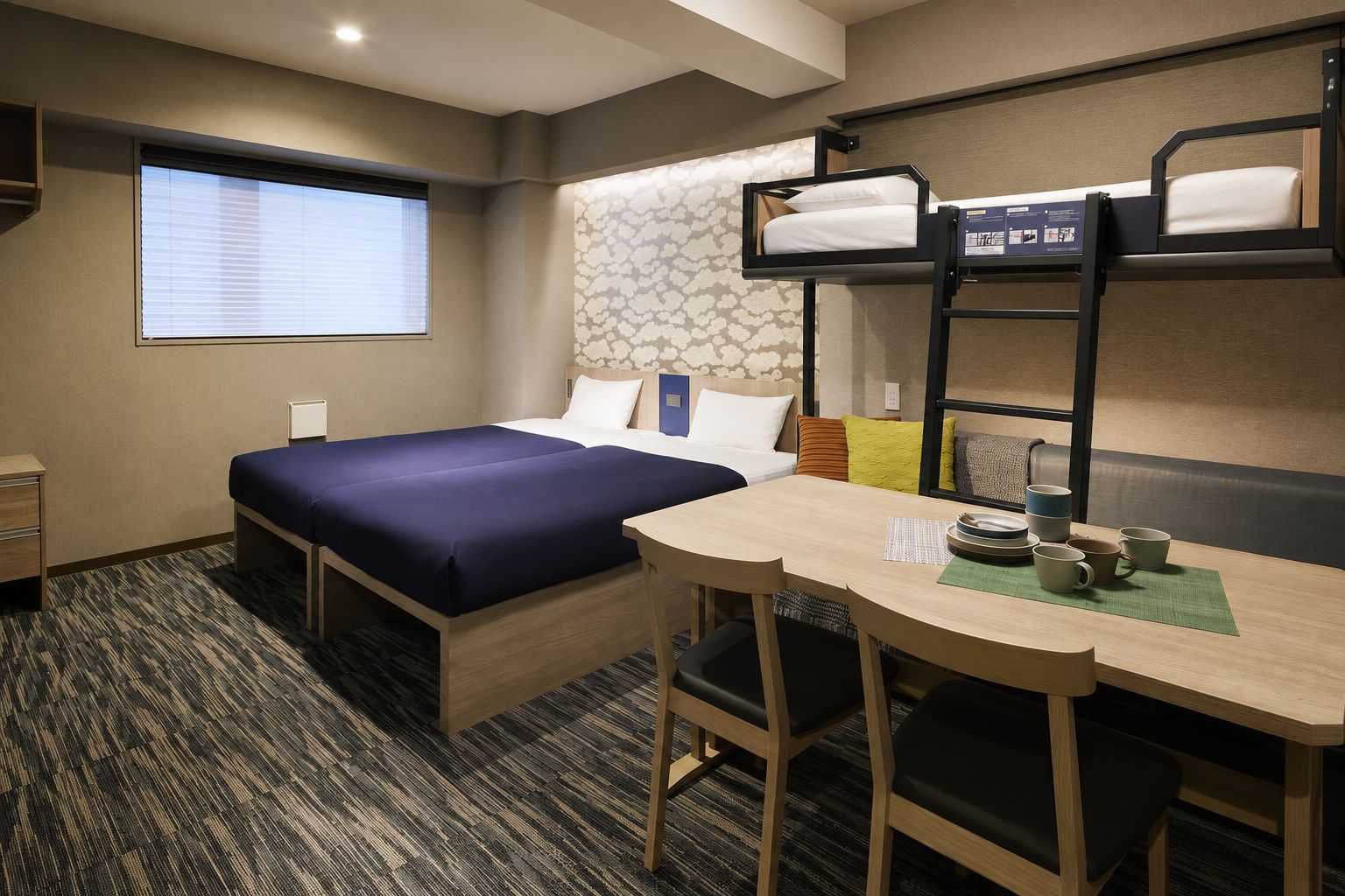 Ein Hotelzimmer mit Etagenbett, einem Einzelbett, einem Schreibtisch mit Stühlen und einem Fenster mit Jalousien.