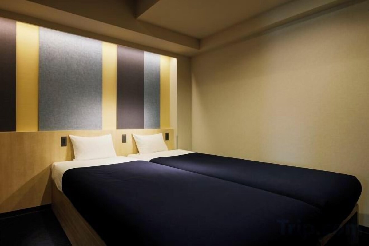 Habitación de hotel con una cama, dos almohadas, cabecera de rayas verticales y una pared lisa.
