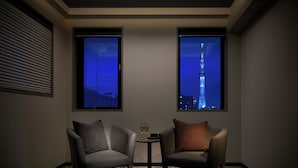 Habitación de hotel con dos sillones, una mesita con dos copas de champán y dos ventanas que ofrecen vistas a un horizonte urbano y una torre.